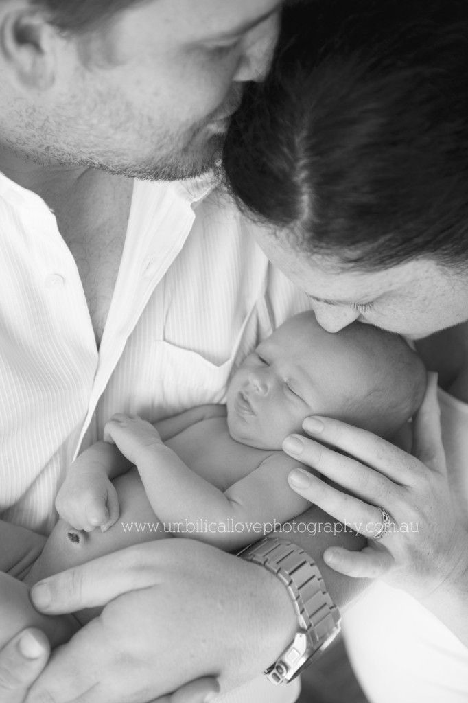 a photo of a brand new baby dad holding baby close while mum kisses babys head and dad kisses mums head