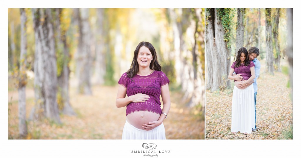 Happy mum to be standing in trees holding belly with husband in the woods looking at the camera