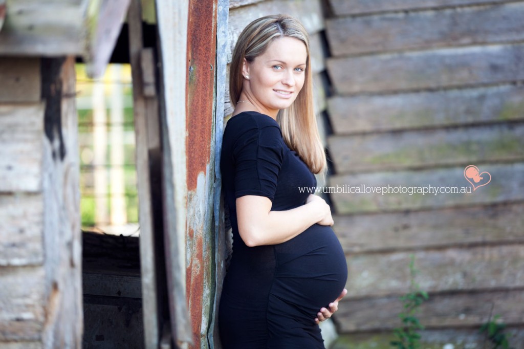 lady who is pregnant holding her belly leaning on an old shed looking at the camera