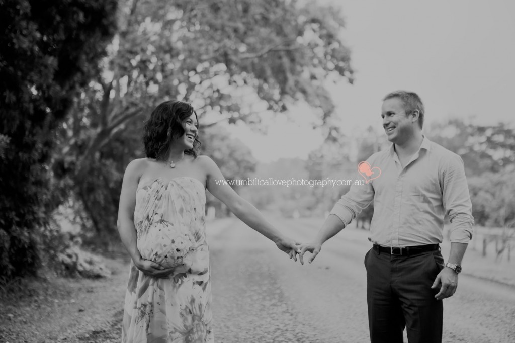 pregnant mum at 35 weeks gestation holding her belly with one hand holding her husbands hand with the other & looking at her husband with a contented smile