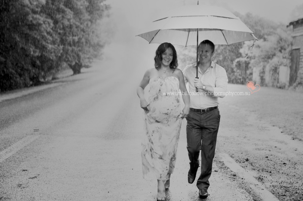 a husband & Pregnant wife walking in the rain smiling at the camera holding a umbrella for their maternity photoshoot