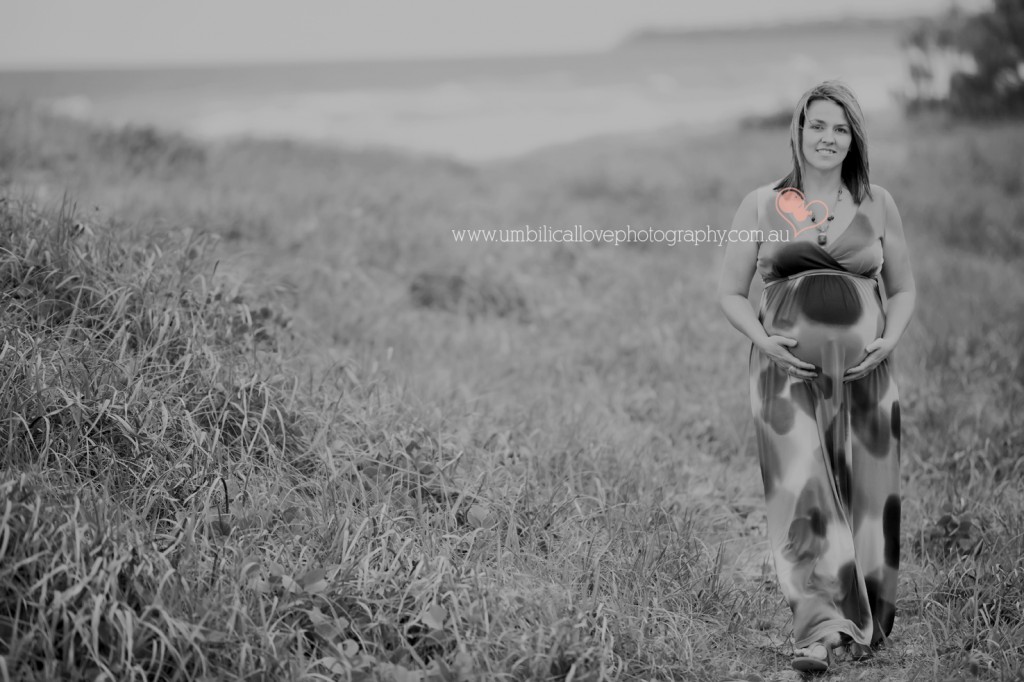 Pregnant lady walking at the beach looking at the camera holding her belly with wind through her hair