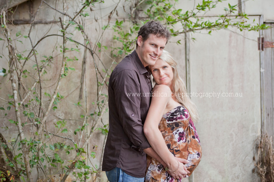Pregnant mum leaning on expecting dad smiling looking at the camera, with rustic shed in background