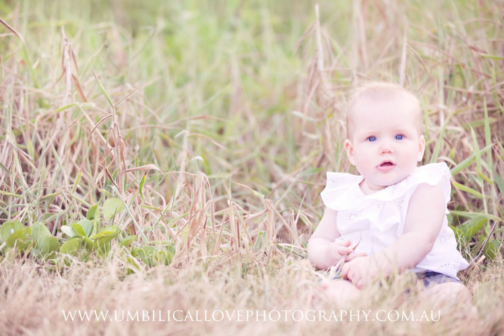 10 month old baby sitting up looking at camera sitting in a grassy patch at mooloolaba beach