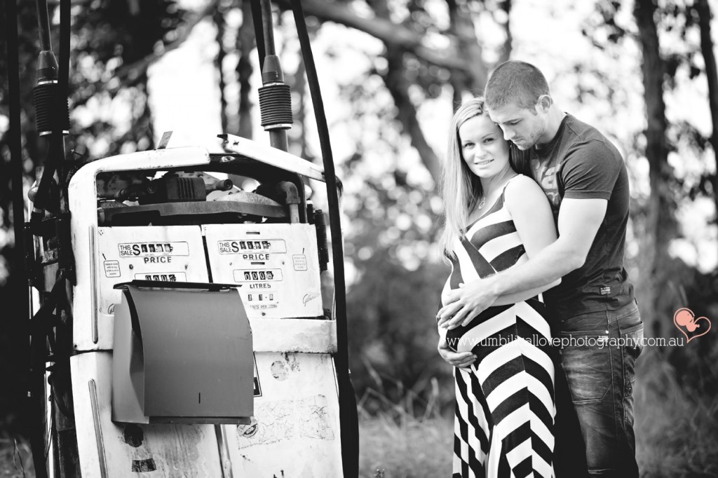 pregnant lady looking at the camera while her husband cuddles her standing next to a vintage petrol pump
