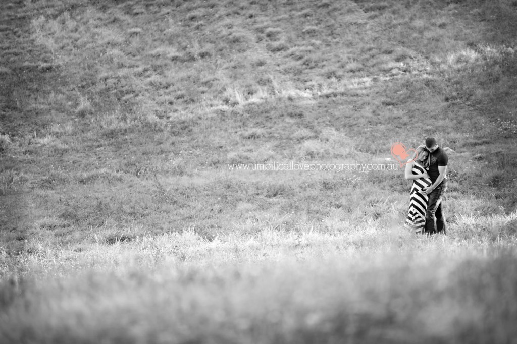 stunning light streaming across a field a pregnant woman & her husband are standing in holding her unborn child