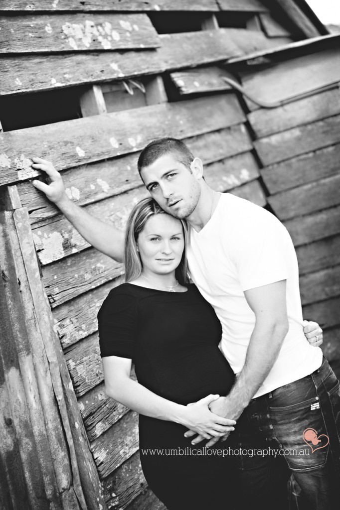 husband leaning on the old shed holding his pregnant wifes belly looking at the camera