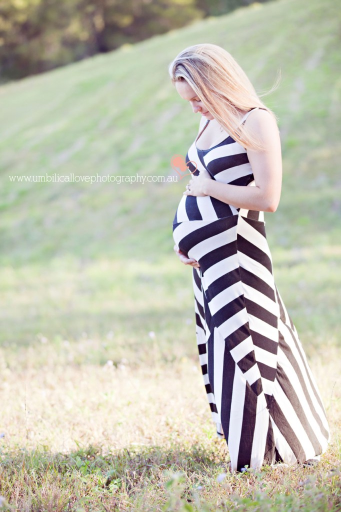 heavily pregnant lady looking down at her belly holding it, standing in a slashed field on the Sunshine Coast